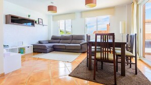 Una sala moderna con un sofá seccional, una mesa de comedor con sillas, un televisor en un estante y ventanas grandes con vistas al exterior.