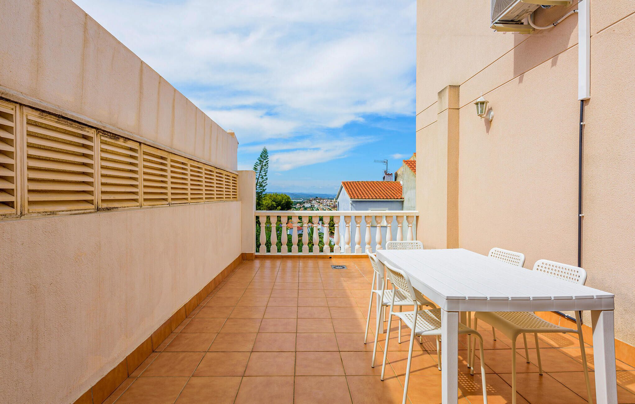 Uma pequena varanda com uma mesa e cadeiras brancas, vista para uma cidade, e um edifício com telhado de telhas vermelhas ao fundo.