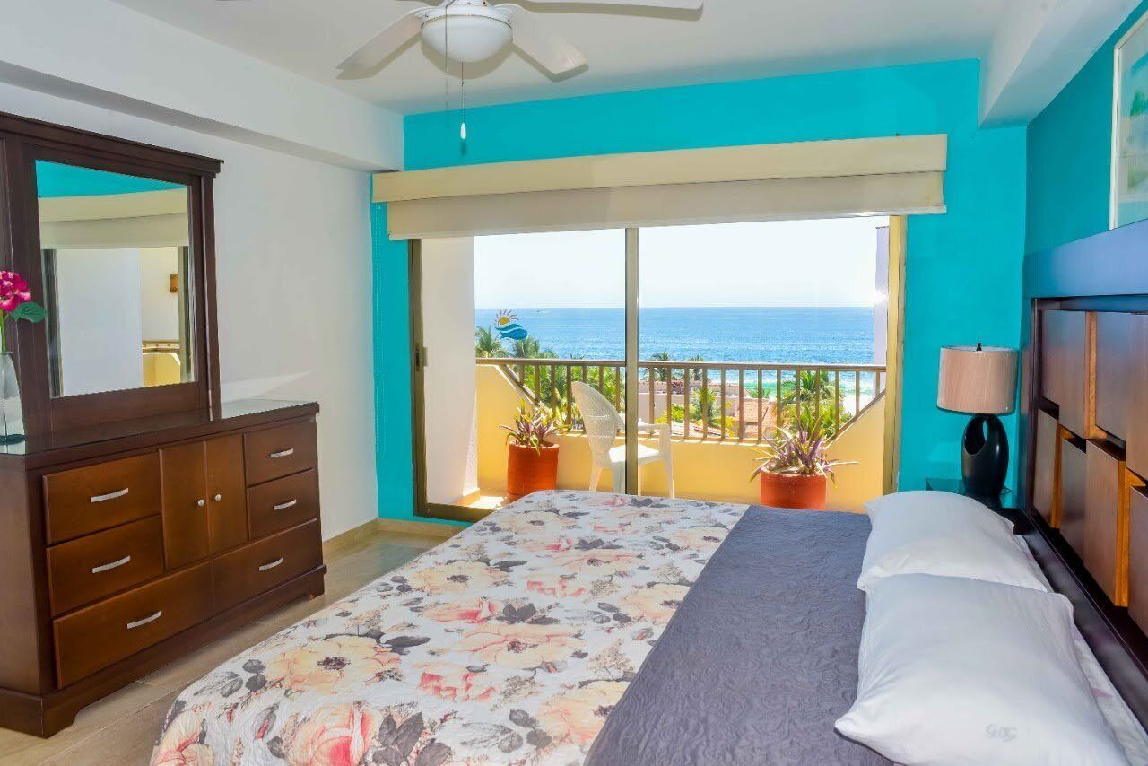 A bedroom with a bed, a dresser, a balcony with plants, and a view of the sea.