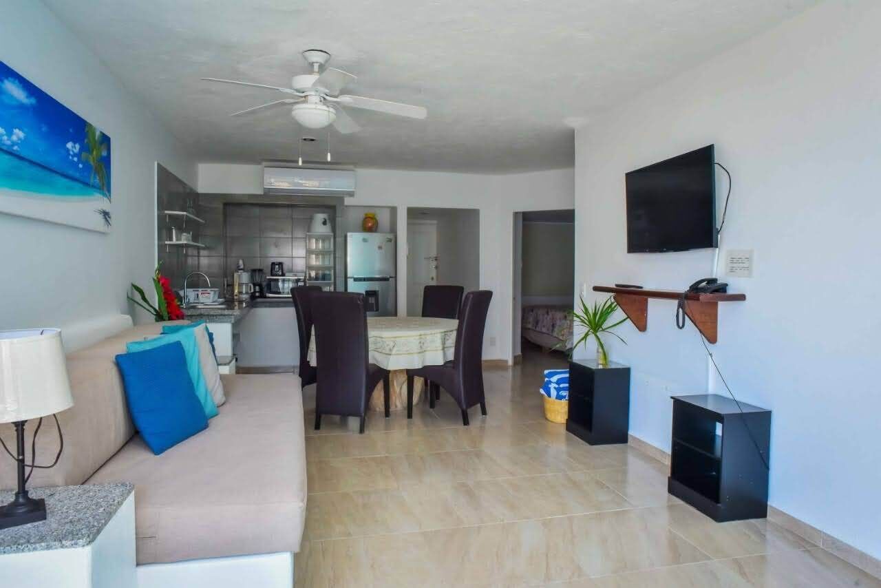 A modern living room with a sofa, a dining table with chairs, a ceiling fan, and a TV mounted on the wall.
