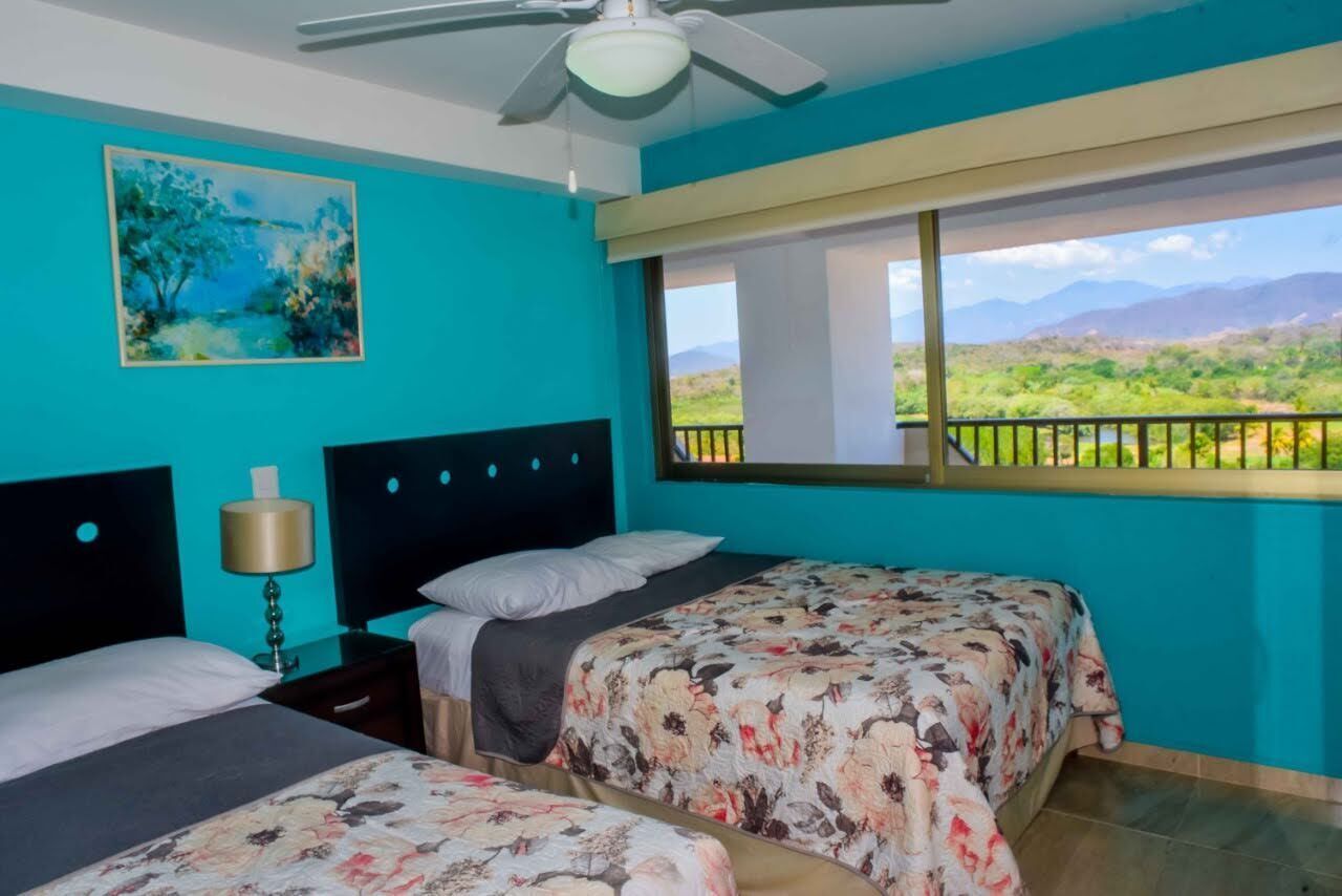 A room with two beds, a ceiling fan, a painting, and a window with a view of greenery and mountains.
