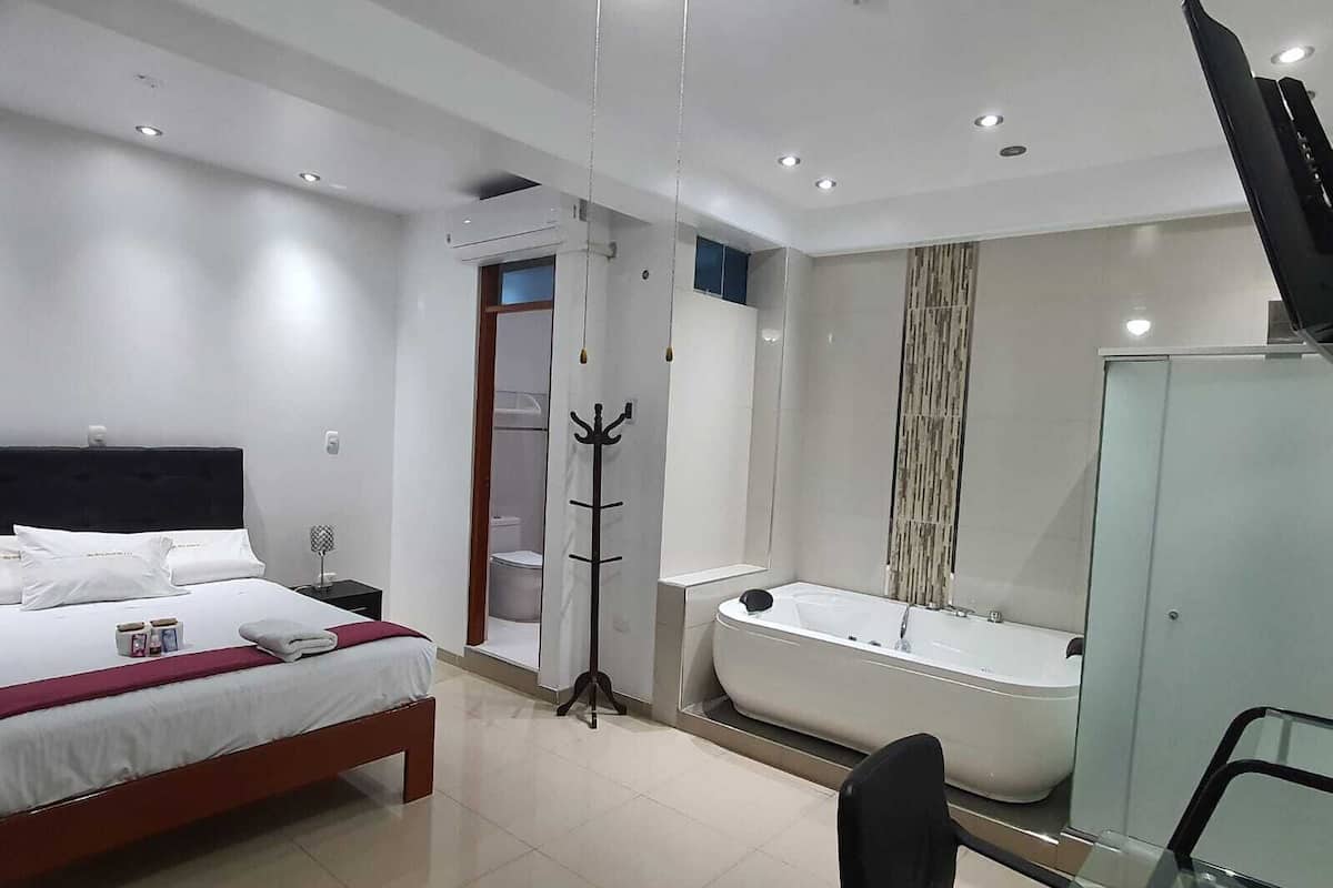 A modern hotel room with a bed, a desk, a chair, a coat rack, a bathroom with a bathtub, and a television.