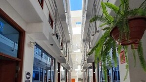 A long, narrow hallway with a high ceiling, flanked by glass doors and windows, leading to an open area with a view of the sky. - Hotel Golden Sky (Pariñas)