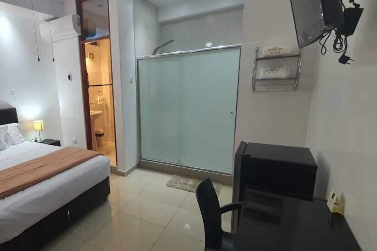 A hotel room with a bed, bedside table, lamp, wardrobe, and a desk with a chair.