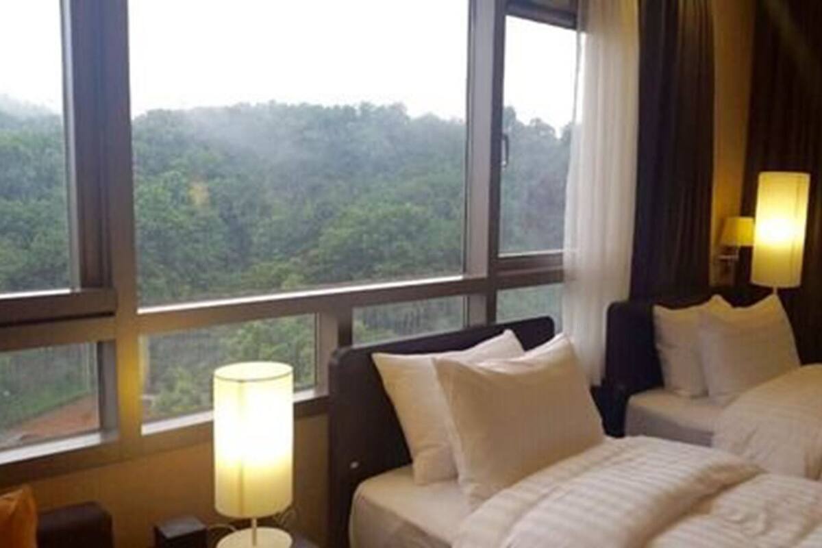 Une chambre d’hôtel avec deux lits, une table de chevet, une lampe et une vue sur une zone boisée.