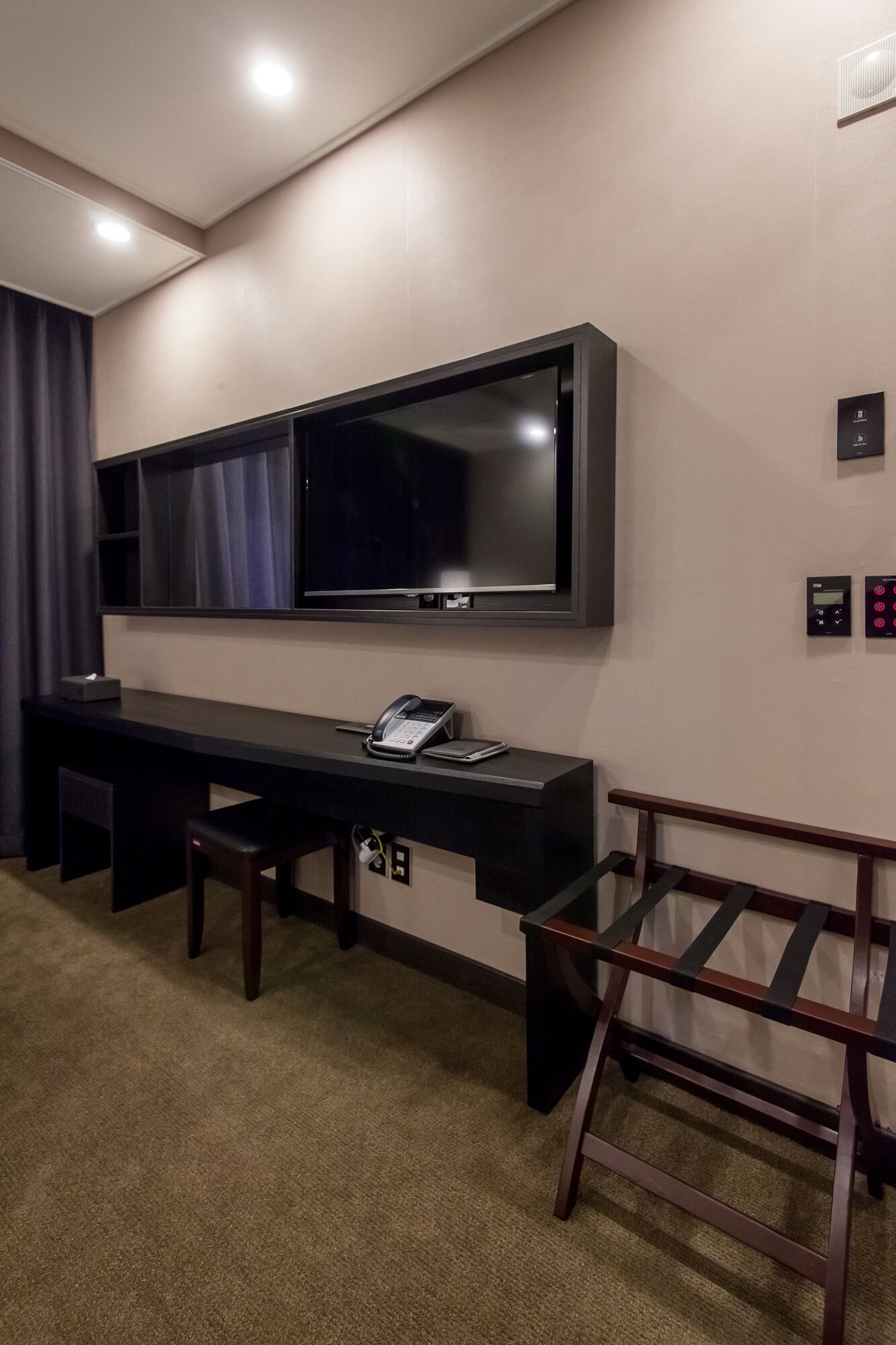 A modern hotel room with a large flat-screen TV mounted on the wall, a desk with a telephone and a small box, a bench, and a wooden chair.