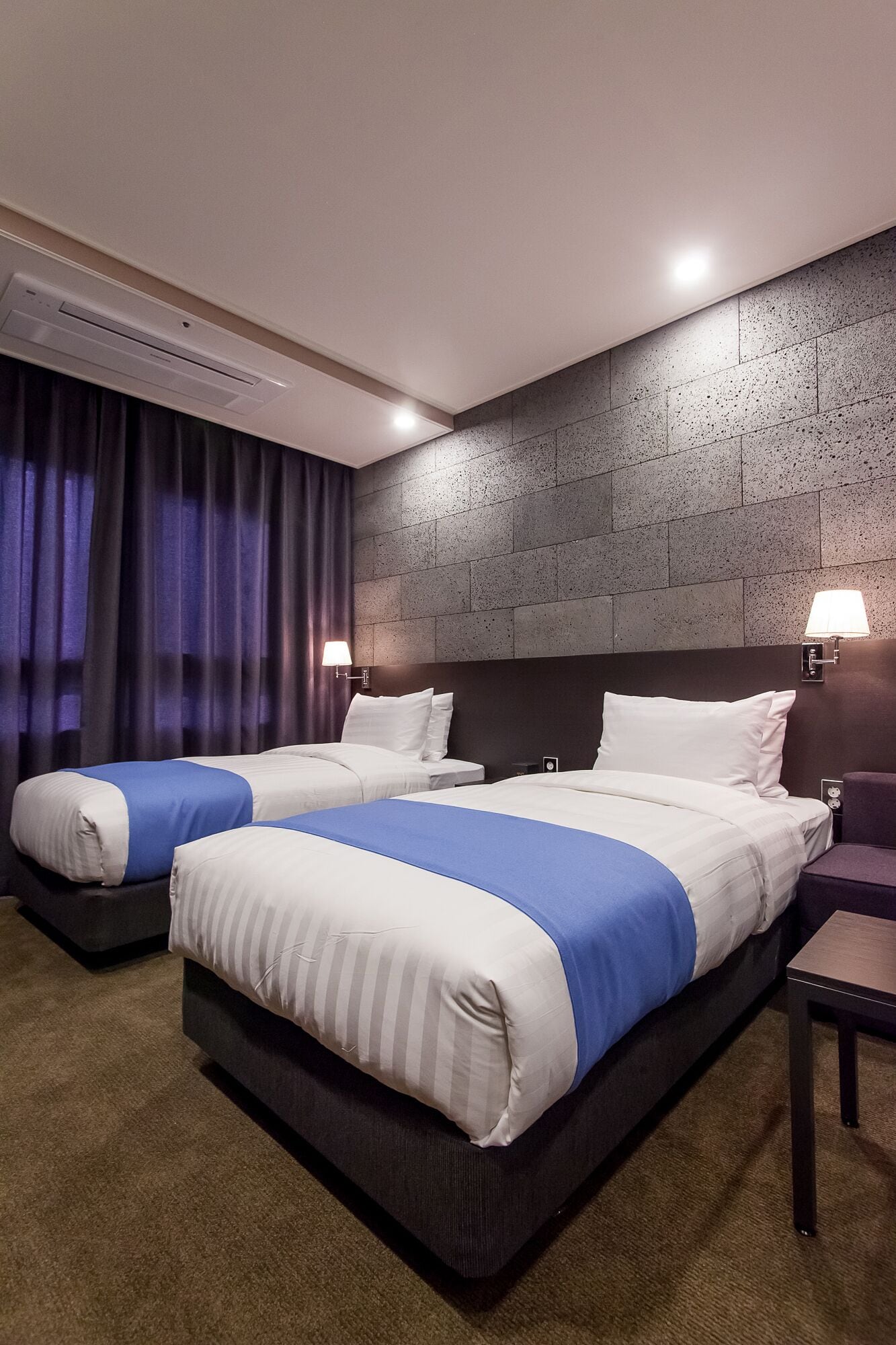 A hotel room with two beds, a dark headboard, a wall-mounted lamp, and a window with curtains.