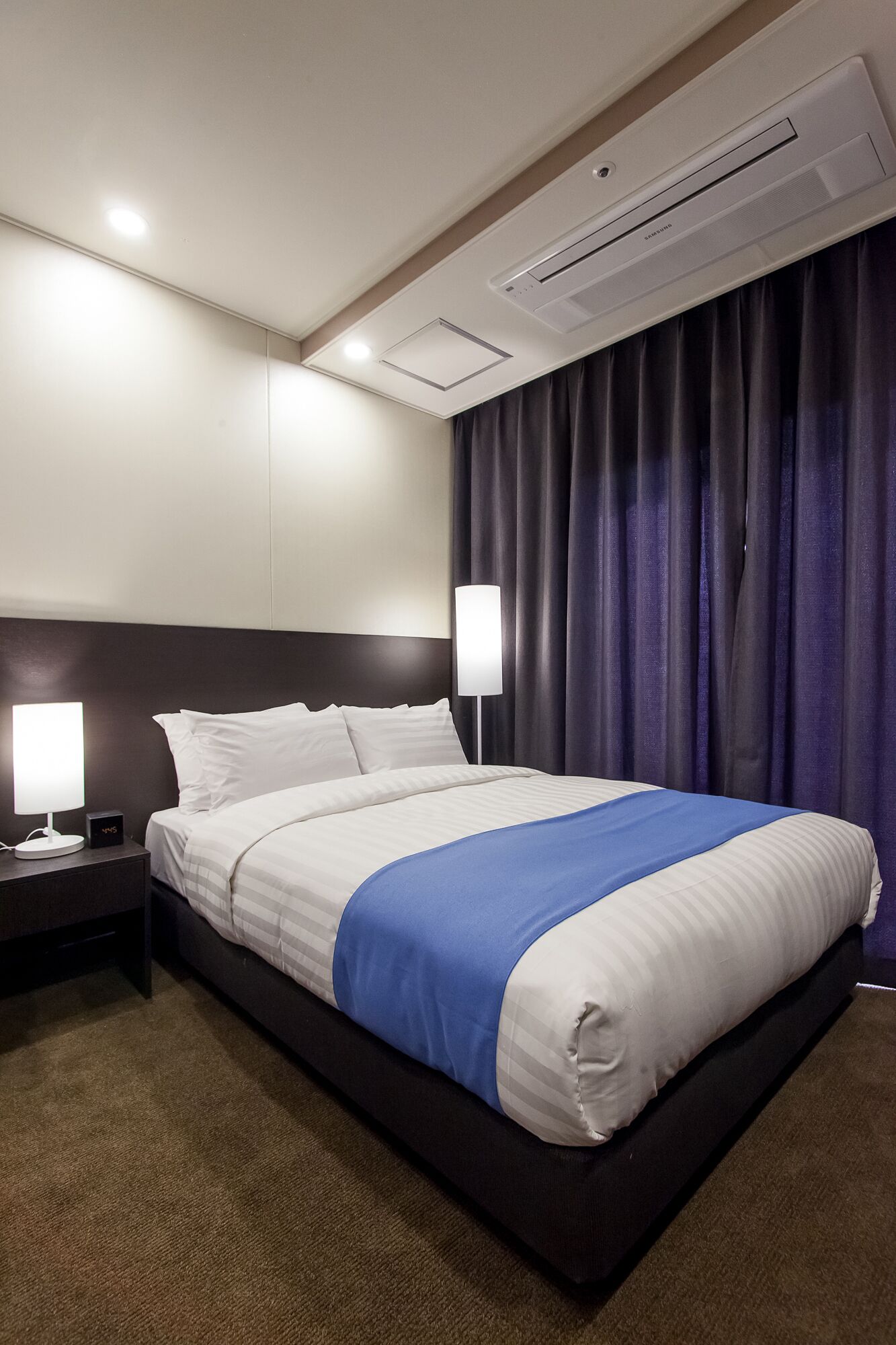 A hotel room with a bed, nightstands, a lamp, and an air conditioning unit.