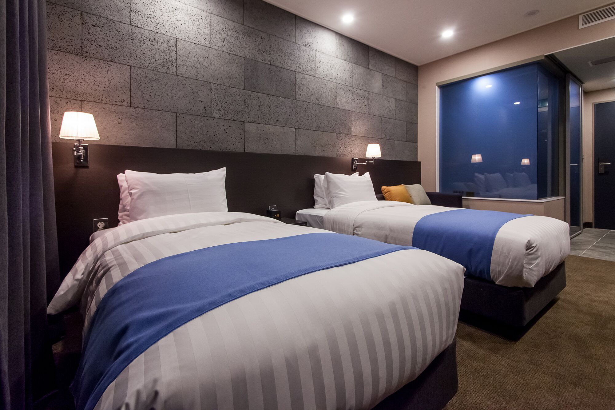 Une chambre d’hôtel avec deux lits, chacun agrémenté de draps blancs et de touches de couleur bleue. Des lampes murales sont fixées de chaque côté des lits. La chambre est dotée d’une tête de lit sombre et d’une grande fenêtre offrant une vue sur l’extérieur.