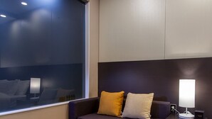 Una habitación de hotel moderna con un ventanal grande, un sofá con cojines, una mesita y una lámpara de noche.