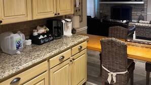 Una cocina con armarios de madera, encimera de granito y cafetera. Se observa una silla de mimbre y una chimenea al fondo.