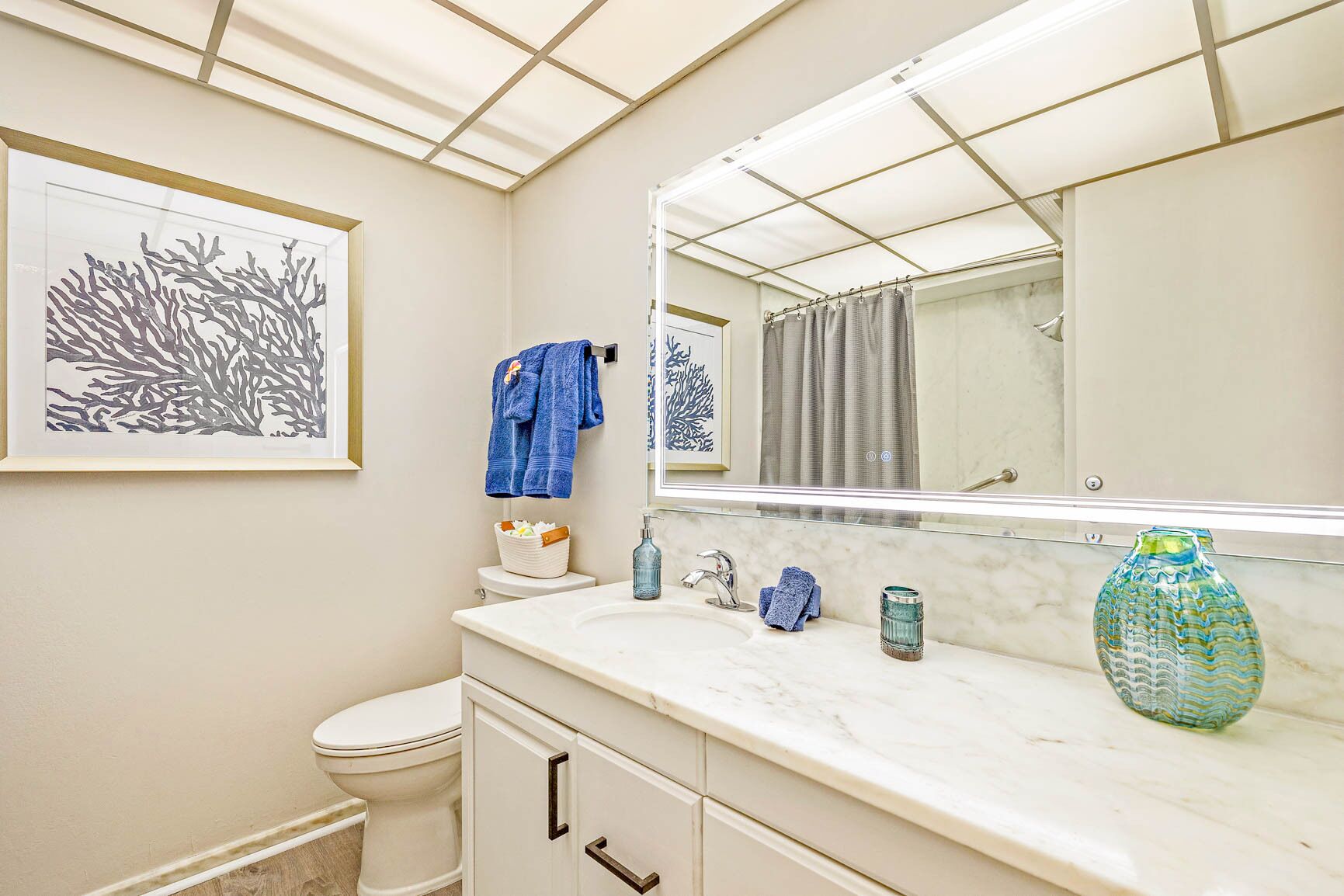 Een badkamer met een wit toilet, een marmeren wastafel, een grote spiegel en een kunstwerk in een lijst aan de muur.