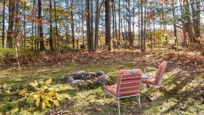 A forested area with a fire pit and two chairs. - Gilford Central Retreat - For all your Vacation Needs! (Gilford)
