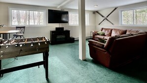 A spacious room with a foosball table, a leather sofa, a flat-screen TV, and large windows. - Gilford Central Retreat - For all your Vacation Needs! (Gilford)