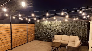 Un espacio al aire libre con patio, con un área de estar acolchada, una mesita y luces colgantes.