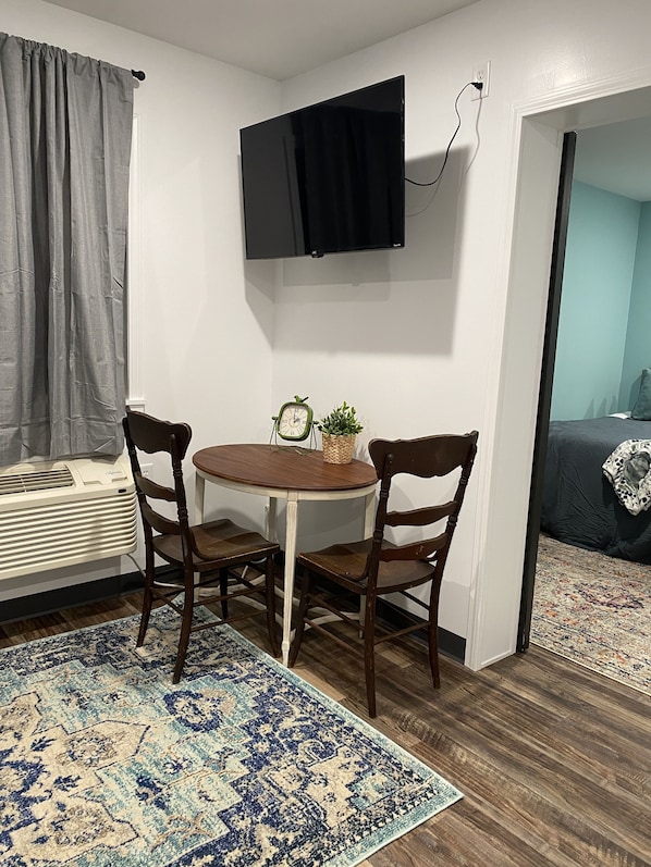 A small room with a round wooden table, two chairs, a television mounted on the wall, a small clock, and a potted plant.