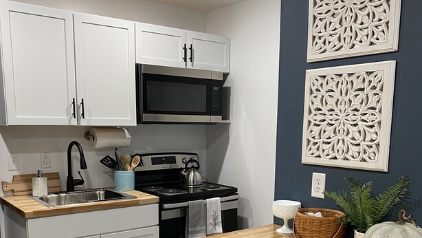 A modern kitchen with white cabinets, a wooden countertop, a stove, and a microwave. There are decorative wall panels and a potted plant on the counter.