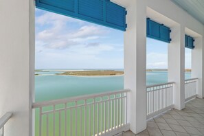 Un balcón cubierto con barandillas blancas y contraventanas azules, con vista a un mar tranquilo y a la costa distante.