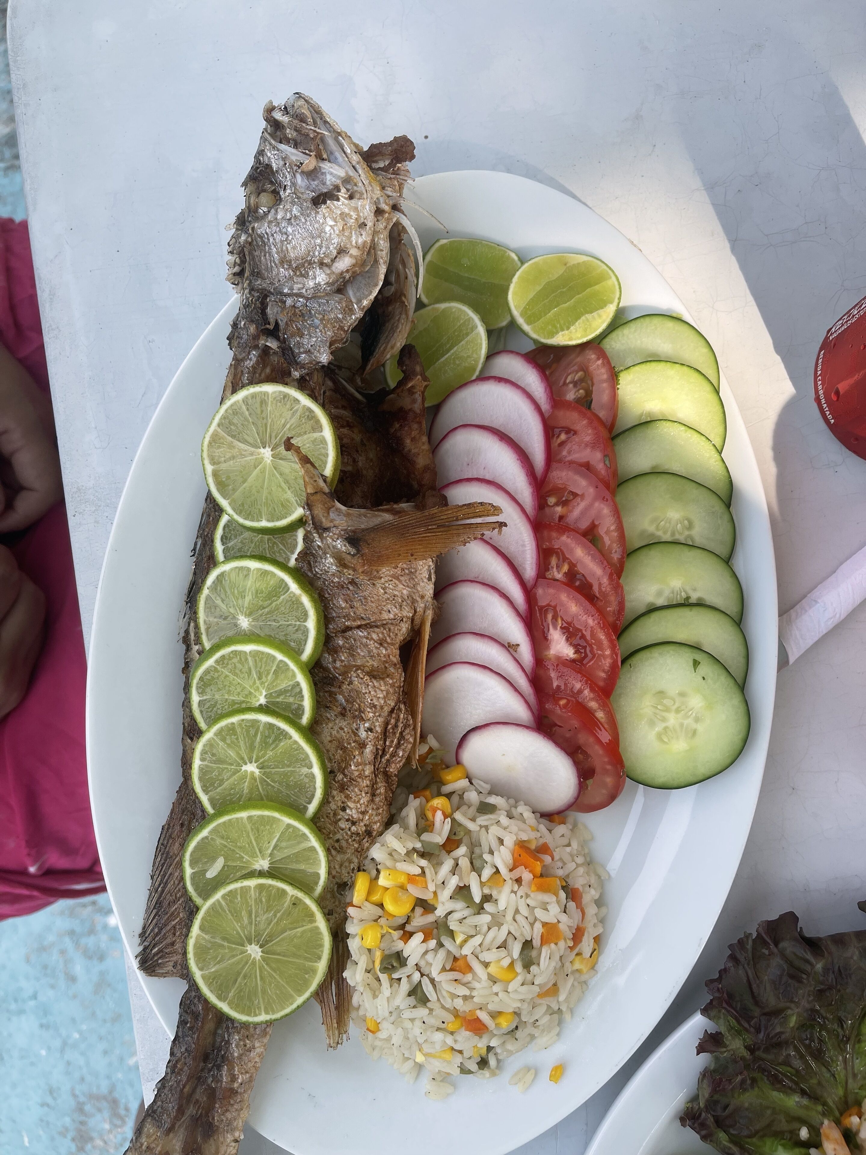 A plate with a whole fish, sliced cucumbers, tomatoes, radishes, and lime slices, accompanied by a side of rice and vegetables.