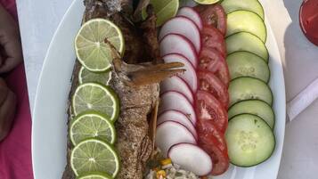 A plate with a whole fish, sliced cucumbers, tomatoes, radishes, and lime slices, accompanied by a side of rice and vegetables.