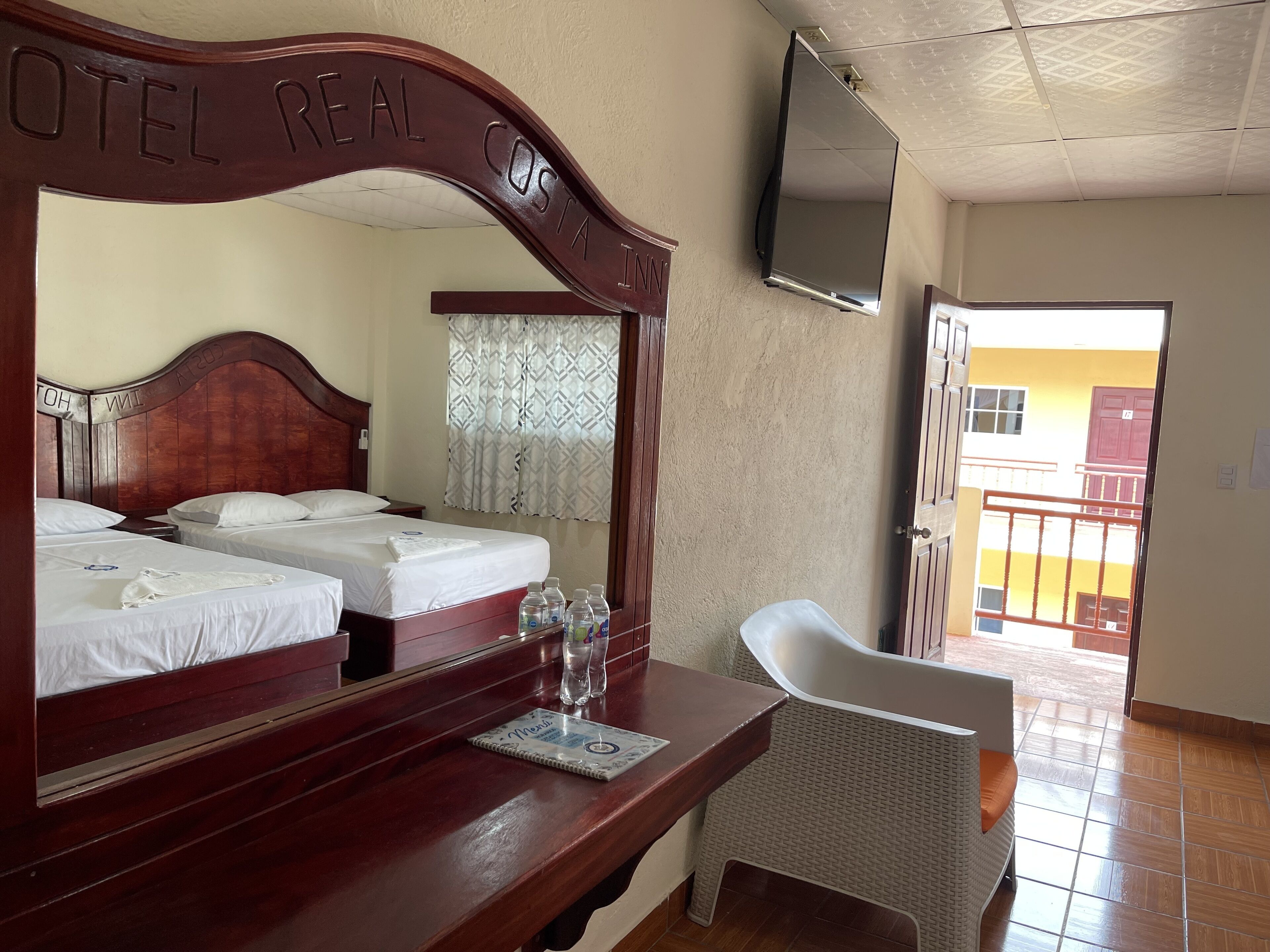 A hotel room with two wooden beds, a desk with a notebook, a chair, and a TV mounted on the wall.