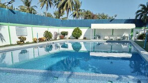 A clear blue swimming pool with a tiled edge, surrounded by a blue and green fence, and palm trees in the background.