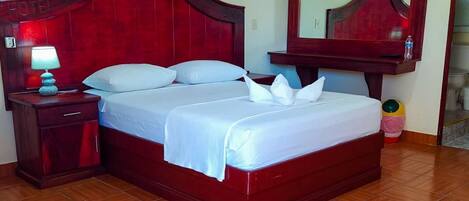 A hotel room with a wooden bed, a nightstand with a lamp, a mirror, and a tiled floor.