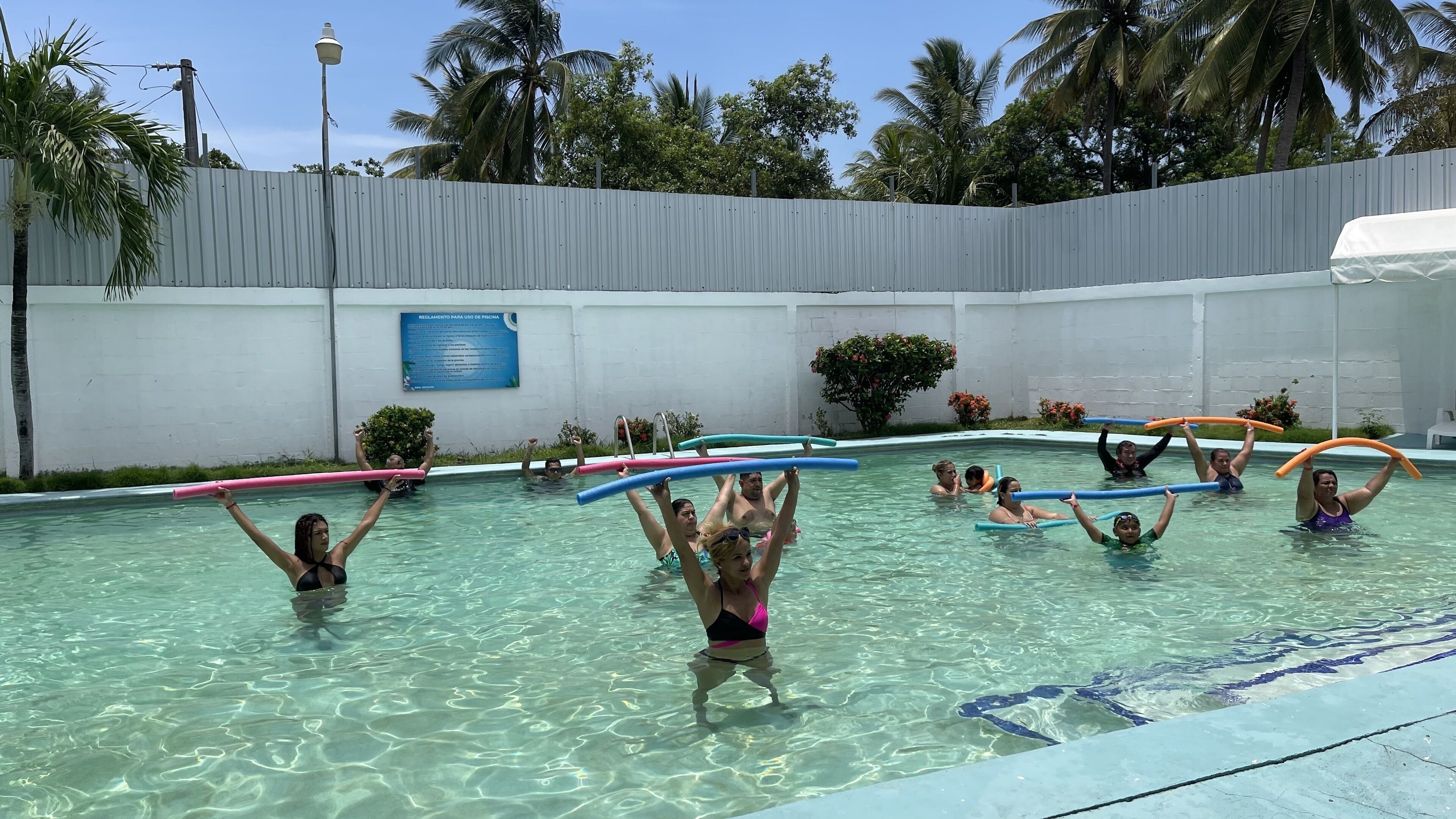 A swimming pool area with clear water, a white wall, and a sign in Spanish.