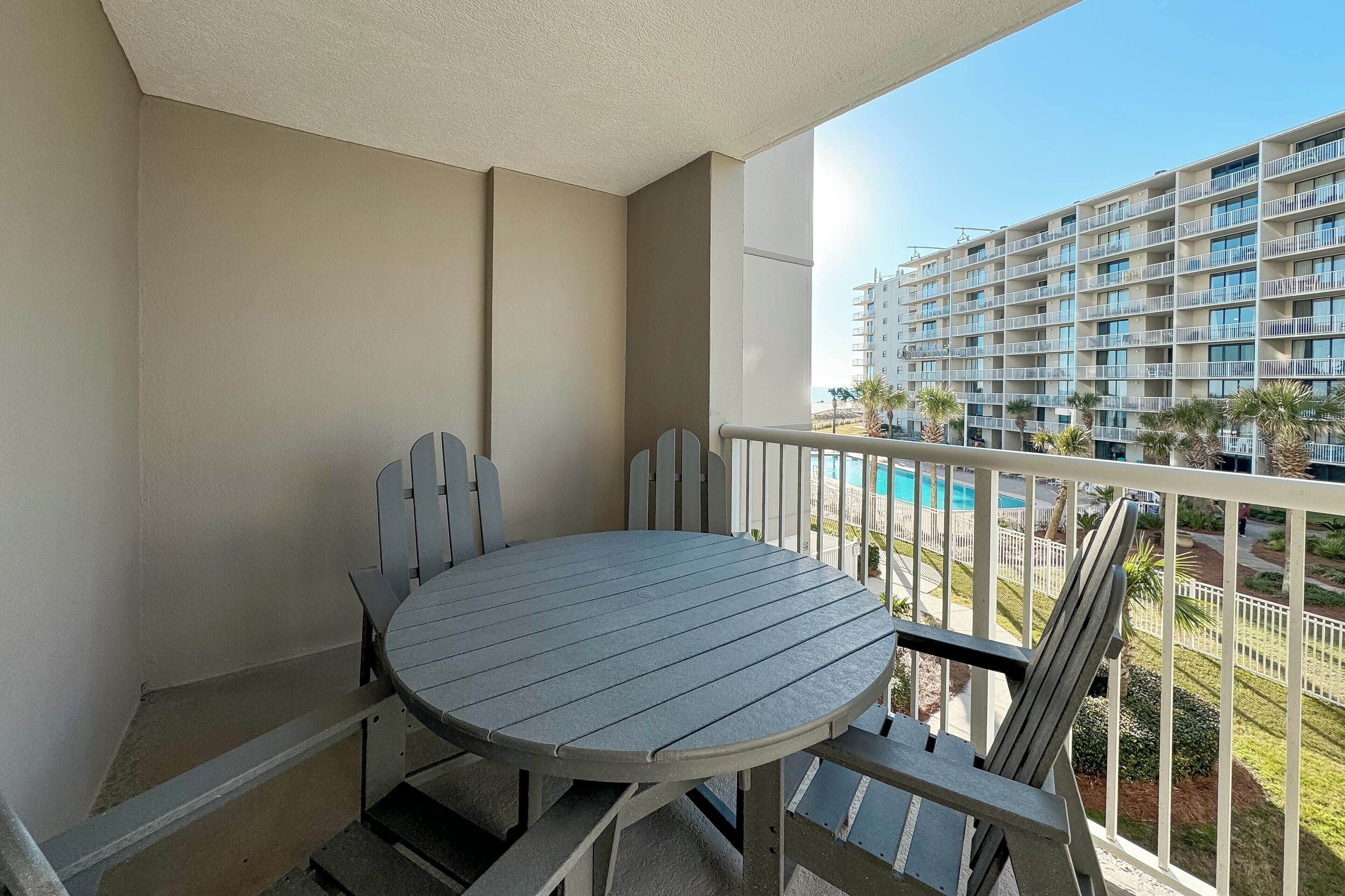 A balcony with a round table and chairs, overlooking a swimming pool and a multi-story building.