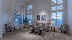A spacious living room with a beige carpet, a central coffee table, a beige sofa with patterned cushions, two armchairs, and large windows with white shutters. - LUX Near Golf & Trails: Pvt POOL/Spa, Wood FP, Game Tables, TVs All BRs, EV Charger (Mesa)