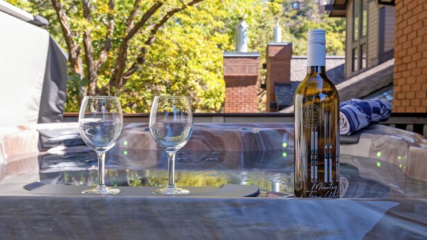 A glass table with two wine glasses and a bottle of wine, set outdoors with a view of trees and buildings in the background.