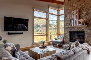 A spacious living room with a stone fireplace, large windows offering a view of a mountainous landscape, a flat-screen TV mounted on the wall, and a comfortable seating area with a sofa and ottoman. - Modern Home with Pool, Gym, Hot Tubs (Silverthorne)