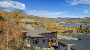 A modern house with a large deck, surrounded by autumn foliage and a view of a lake.