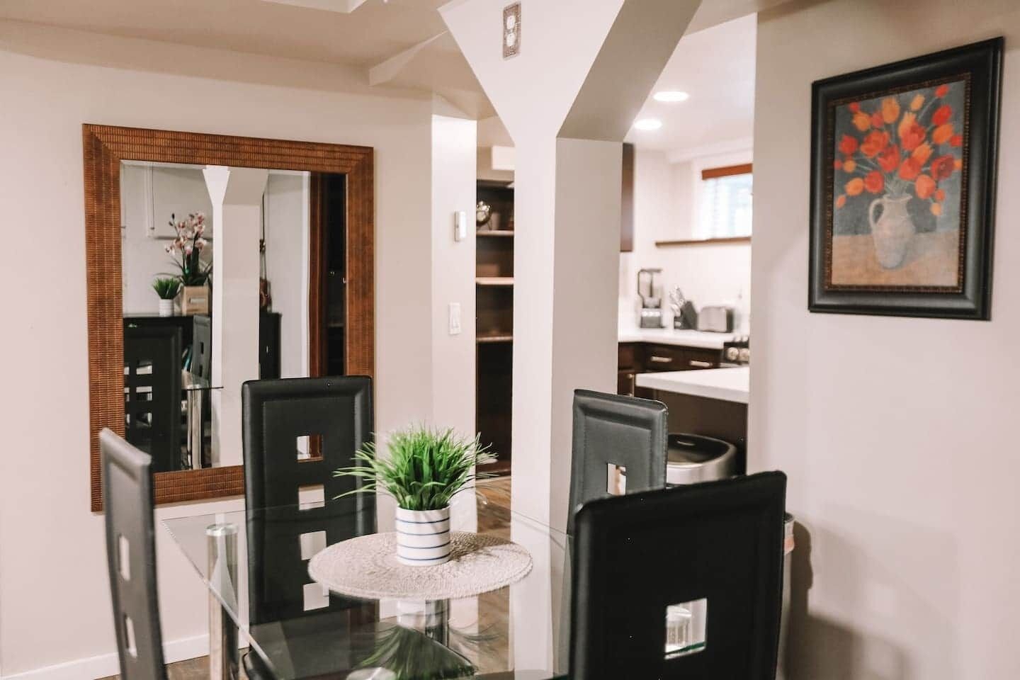 Un coin repas avec une table en verre, des chaises noires, une plante en pot, un miroir et, au fond, une cuisine.