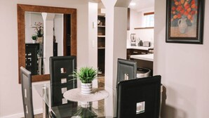 Un coin repas avec une table en verre, des chaises noires, une plante en pot, un miroir et, au fond, une cuisine.
