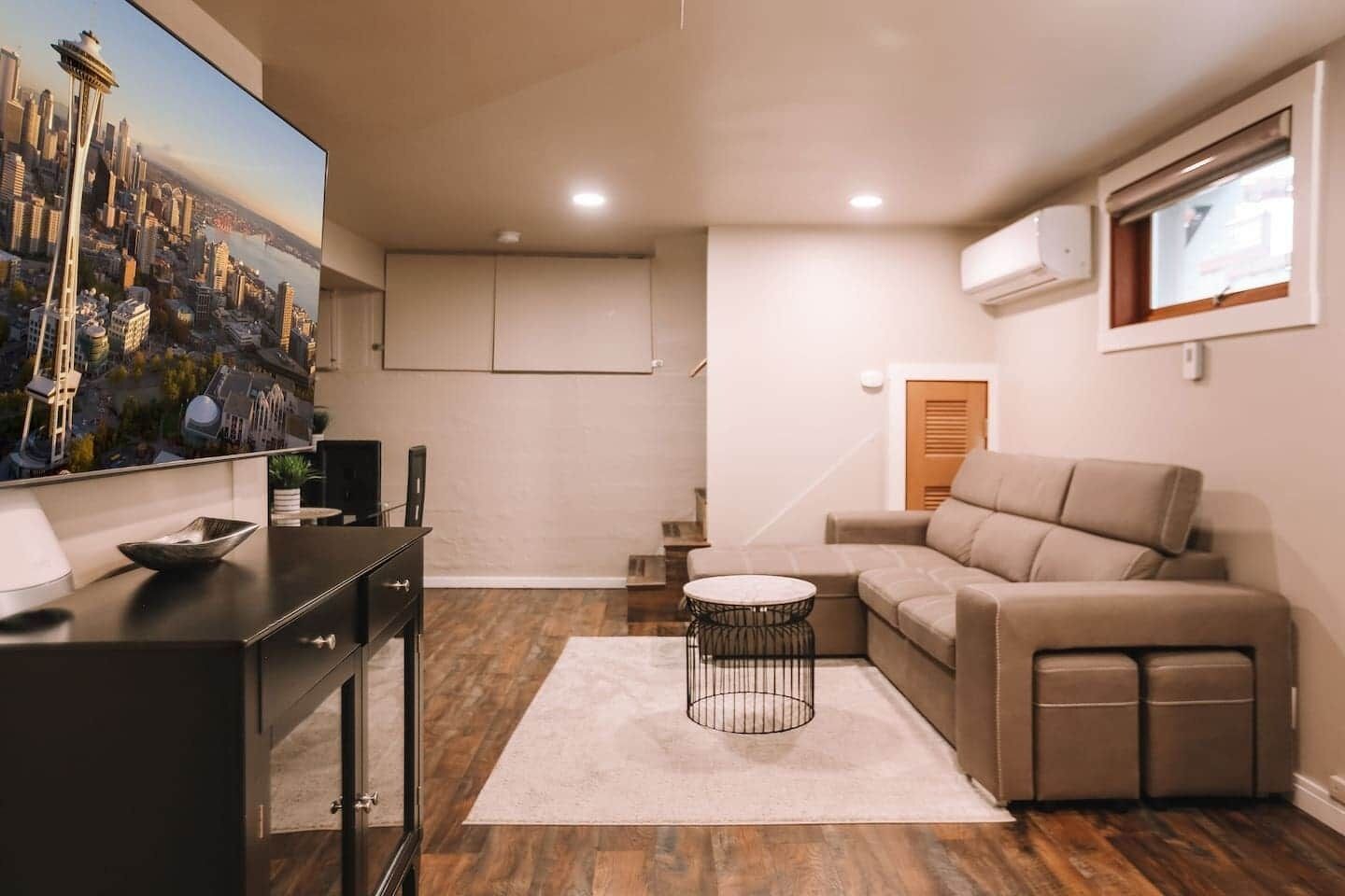 Un salon moderne avec un canapé d’angle, une petite table ronde, un meuble TV noir et un tableau représentant un paysage urbain accroché au mur.