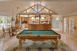 Une table de billard dans une pièce dotée d’un haut plafond et de poutres en bois, un coin repas avec une table et des chaises, et un espace cuisine avec des placards et un plan de travail.