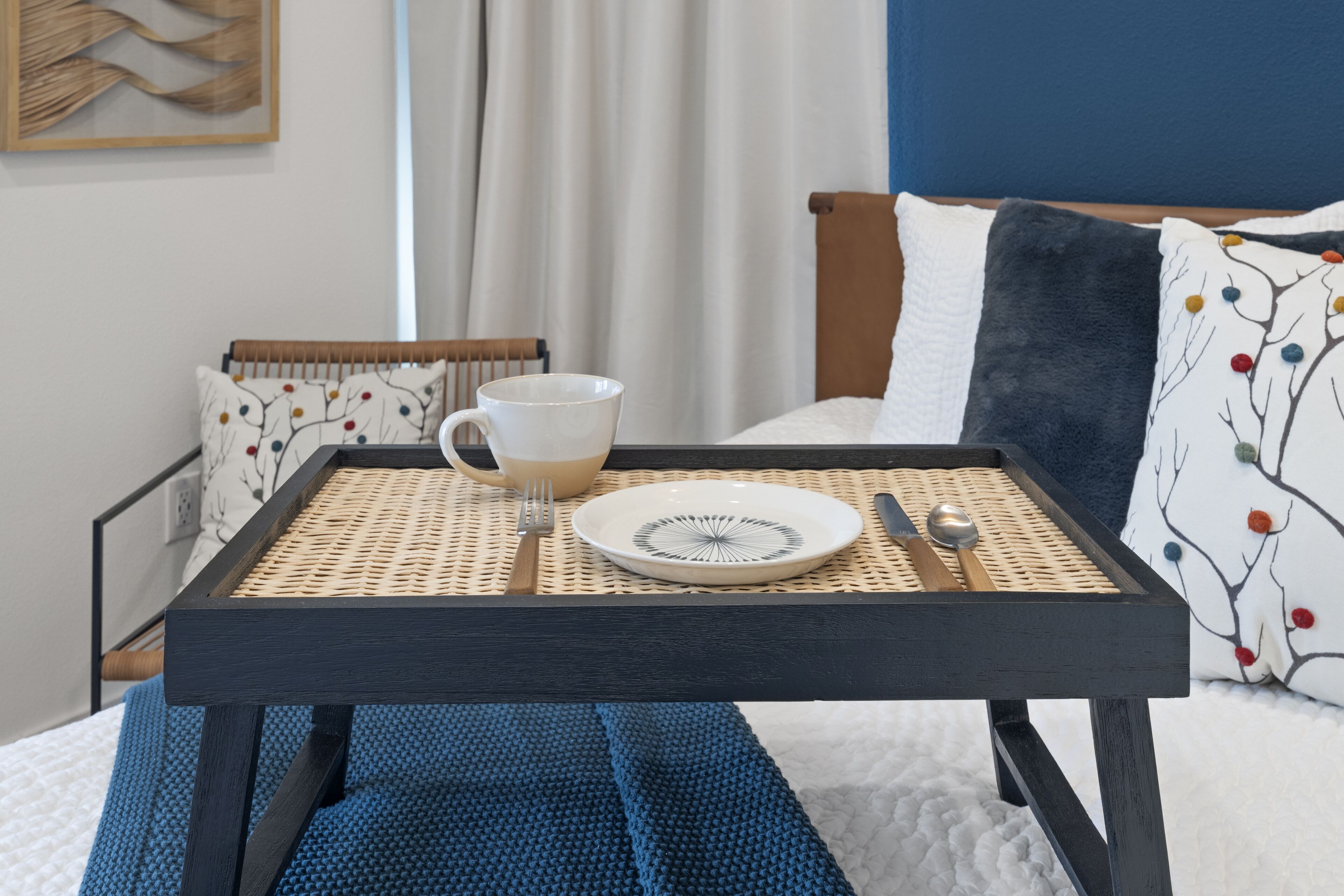 A neatly arranged breakfast table with a plate, cup, and spoon on a woven mat, set against a bed with a blue and white patterned pillow.