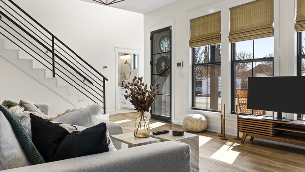 A modern living room with a staircase, a flat-screen TV, and large windows with blinds.