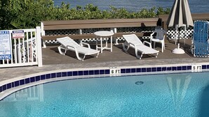 Una piscina con sillones de descanso y sombrillas, rodeada por una terraza de madera y vistas a un lago.
