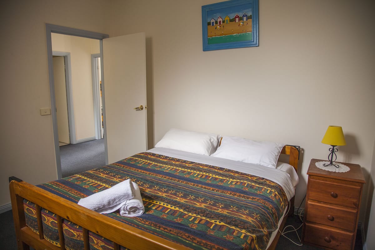 A neatly made bed with a patterned bedspread, a wooden nightstand with a lamp, and a painting on the wall.