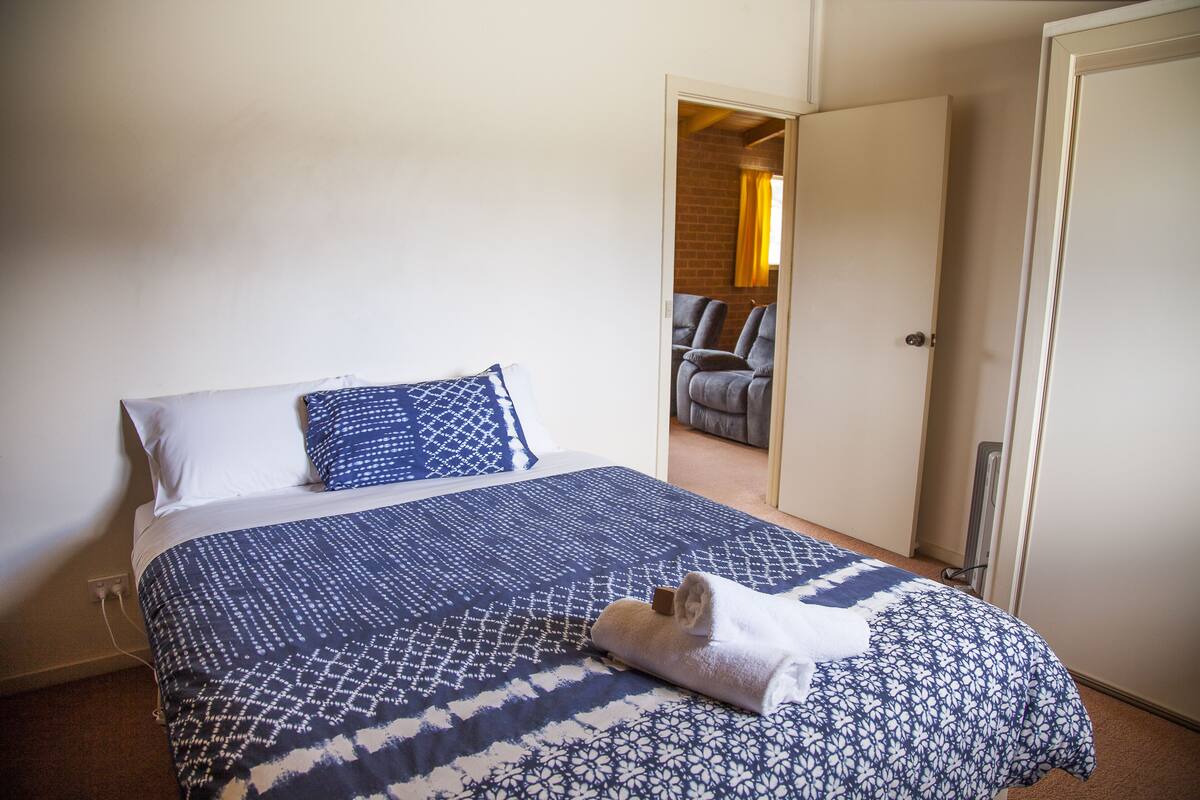 A neatly made bed with a patterned bedspread and pillows, a folded towel on the bed, and a door leading to a room with a brick wall and a chair.