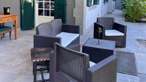 Un área de estar al aire libre con mobiliario de mimbre y una mesa de madera, una puerta con contraventanas verdes y una pared de piedra.