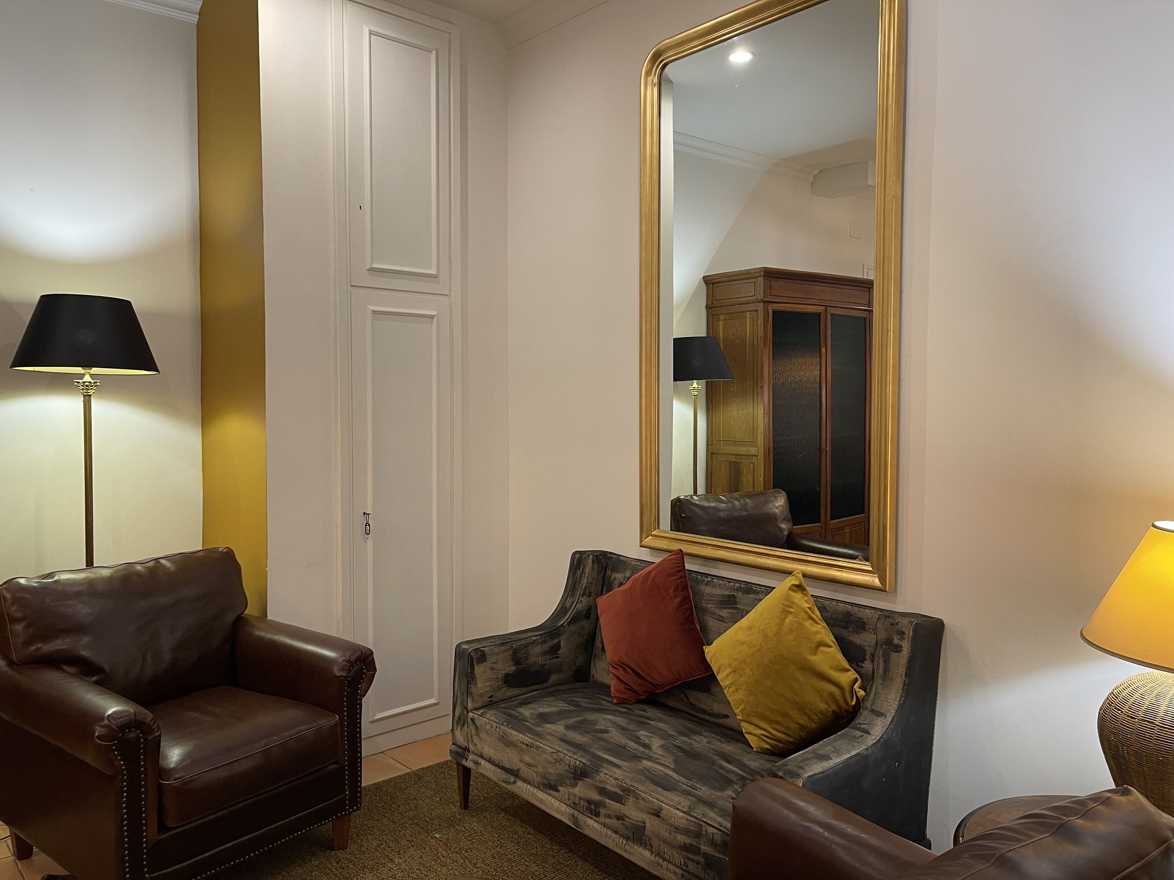 Een kamer met een bruine leren fauteuil, een gestreept bankje met twee kussens, een grote spiegel met gouden lijst en een vloerlamp.