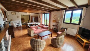 Ein gemütliches Wohnzimmer mit einer Couch, zwei Korbstühlen, einem Couchtisch, einem Fernseher und großen Fenstern mit Blick ins Freie.