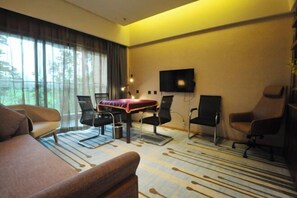 Una habitación de hotel moderna con un sofá, una mesa de comedor con sillas, un televisor de pantalla plana montado en la pared y un gran ventanal con cortinas.