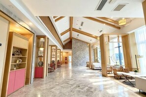Un hall d’hôtel spacieux avec un plafond haut, des poutres en bois et un sol en marbre. On y trouve des coins salon et un comptoir d’accueil.
