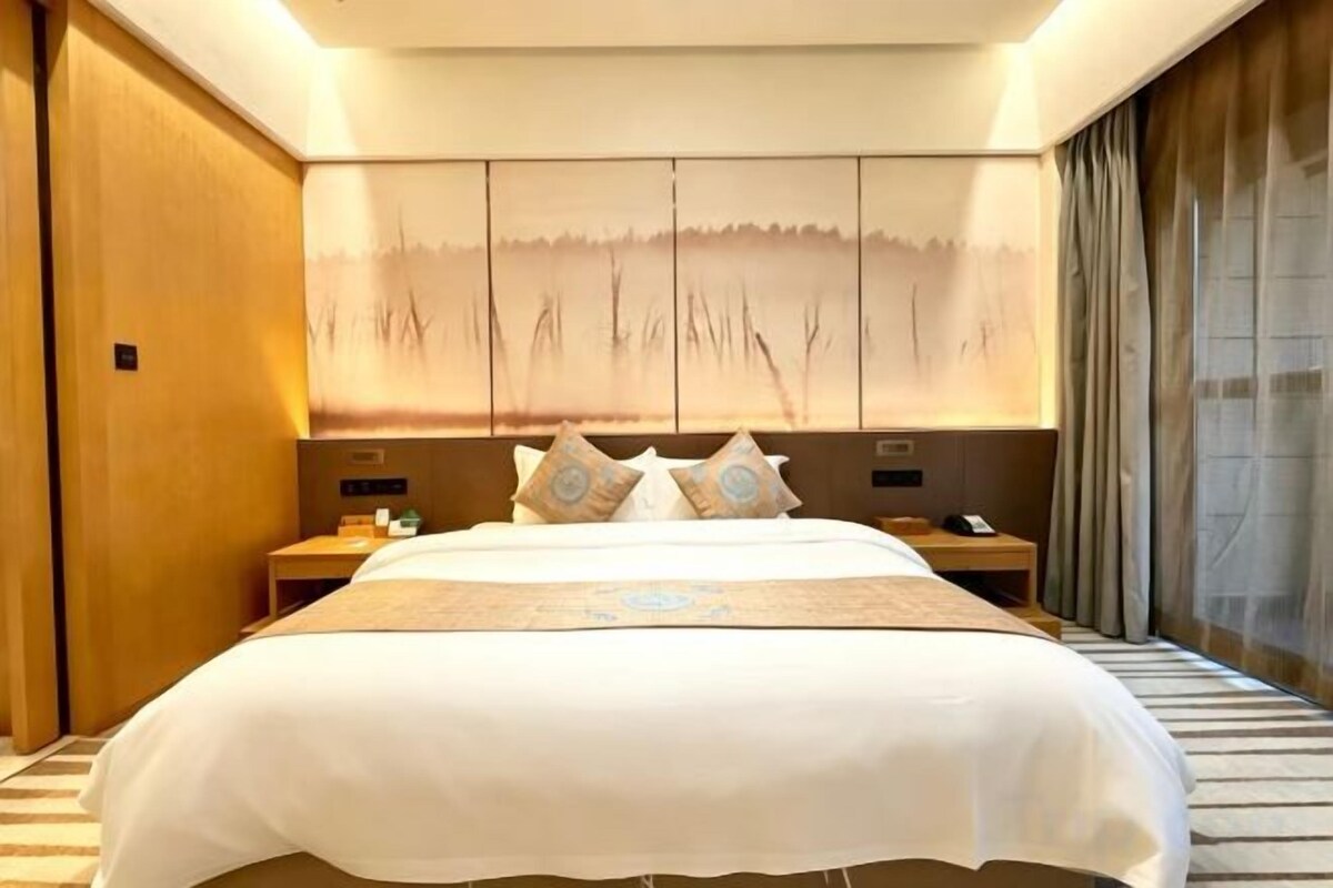 A hotel room with a large bed, a nightstand, a wall-mounted artwork, and a window with curtains.