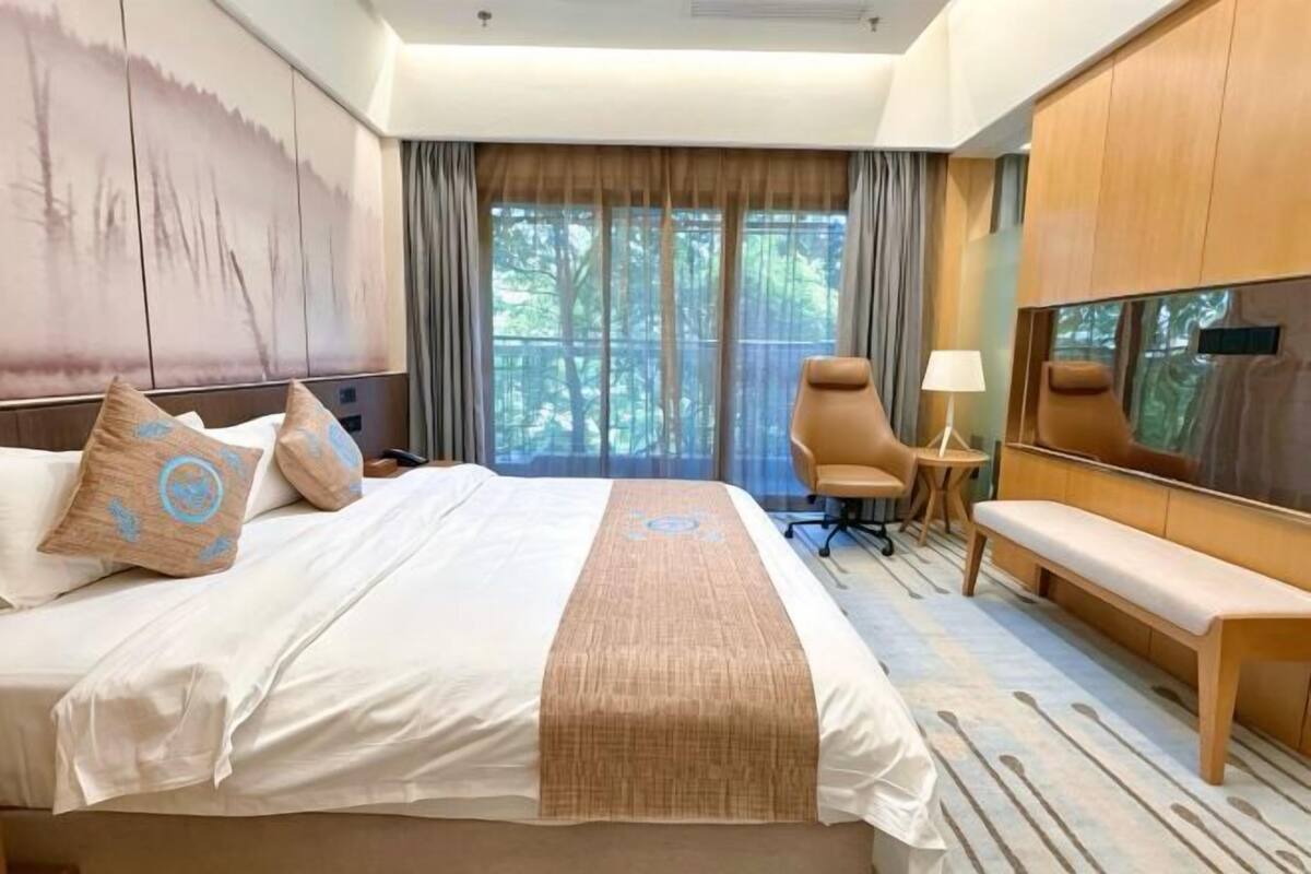 Habitación de hotel con una cama grande, un escritorio, una silla, un banco, televisión y vista a árboles por la ventana.