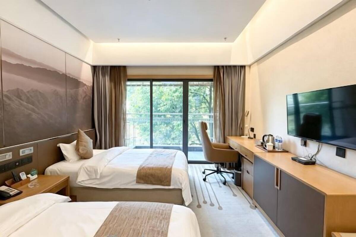 Habitación de hotel con dos camas, un escritorio con televisor, una silla y un ventanal grande.